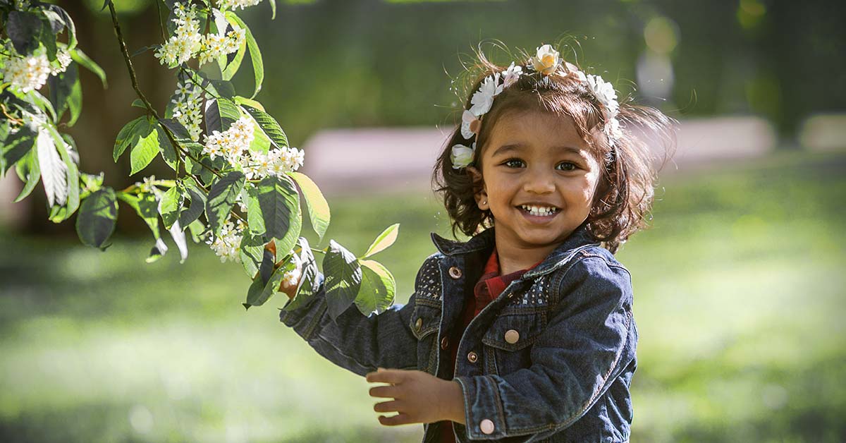 Two-year-old girl with flowers in her hair pulls the leaves of a flowering tree.