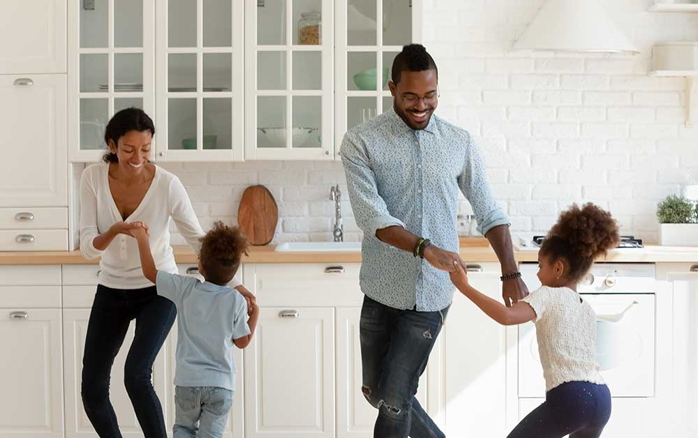 Smiling African American family with a mom, dad, toddler son and daughter dance in their kitchen.